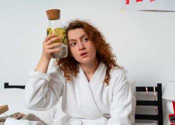 woman holding a bottle of water with lemon and herbs next to a tired person – natural hangover remedies