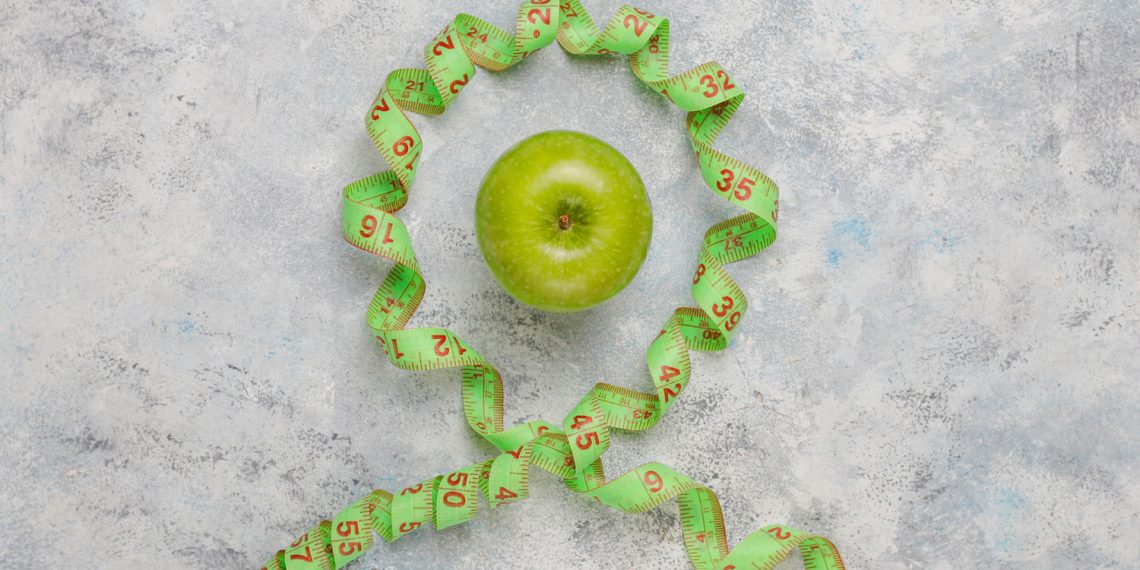 Green apple encircled by measuring tape on concrete background.