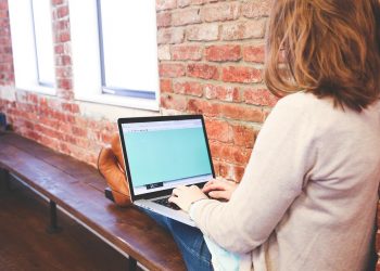 Person using a laptop on a wooden bench against a brick wall.