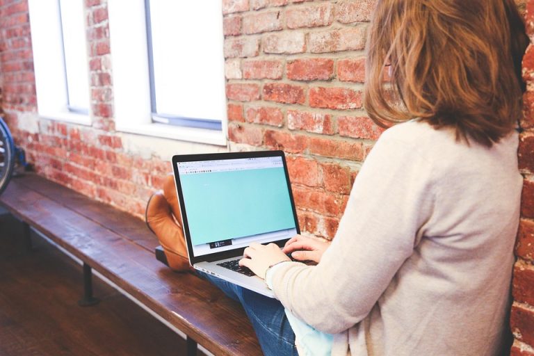 Person using a laptop on a wooden bench against a brick wall.