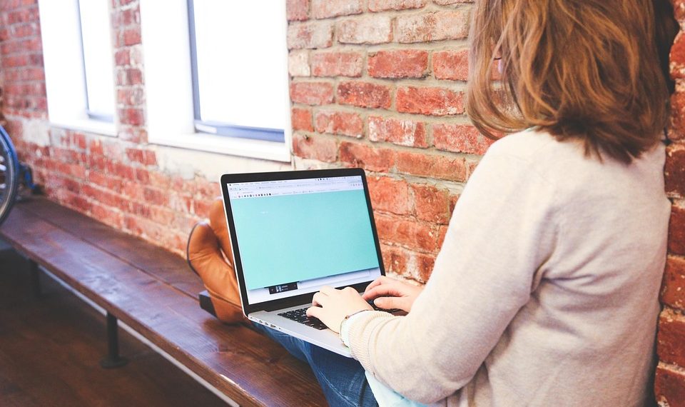 Person using a laptop on a wooden bench against a brick wall.