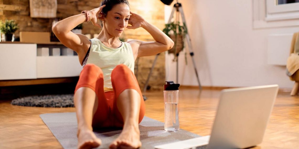 Woman doing sit-ups at home with a laptop for online workout guidance.