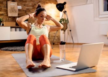 Woman doing sit-ups at home with a laptop for online workout guidance.
