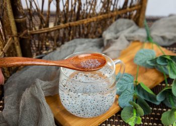 Spoon with chia seeds over glass cup on wooden tray.