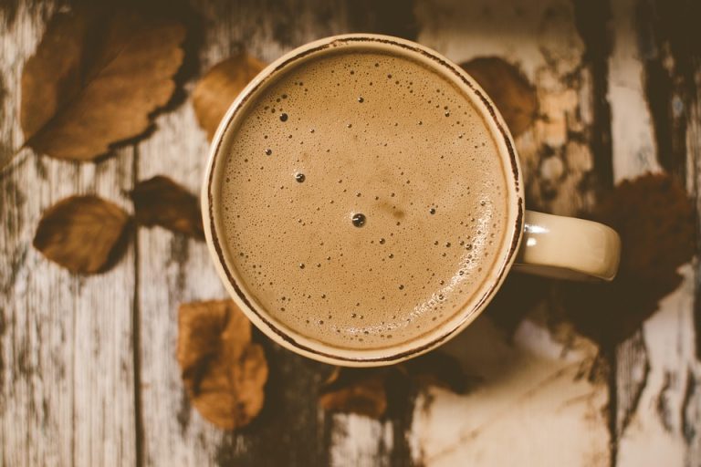 Creamy coffee in a mug with autumn leaves on wooden table.