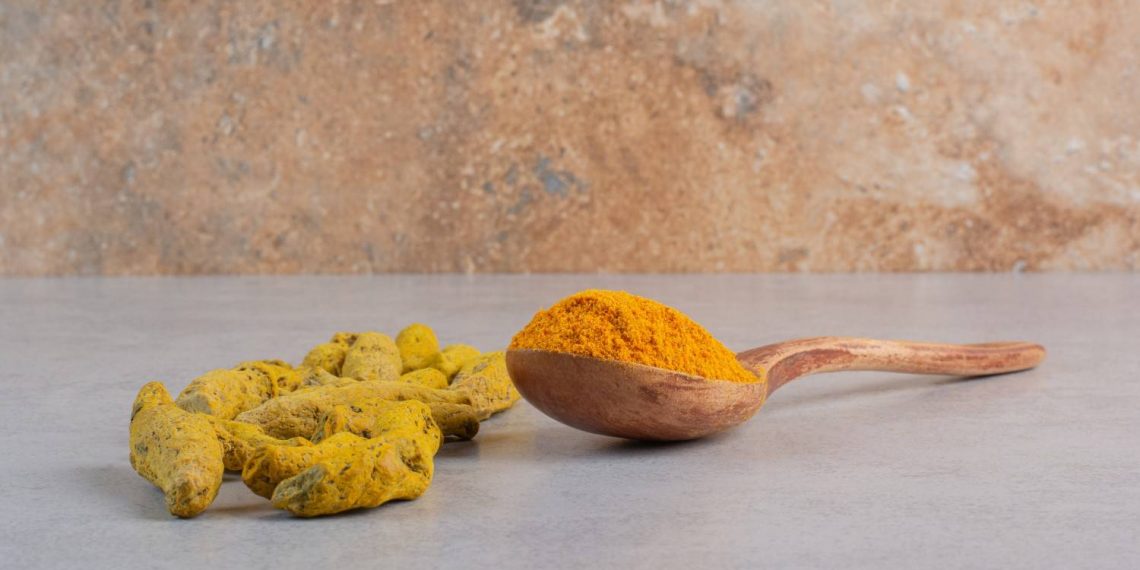 Turmeric roots and powder on a wooden spoon on a marble surface.