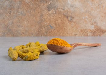 Turmeric roots and powder on a wooden spoon on a marble surface.
