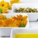 Yellow calendula flowers, dried herbs, and oil in white bowls.