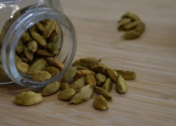 Green cardamom pods spilling from a glass jar on a wooden surface.