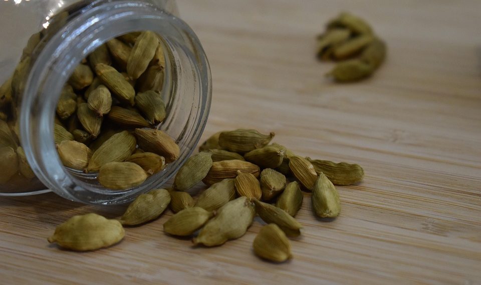 Green cardamom pods spilling from a glass jar on a wooden surface.