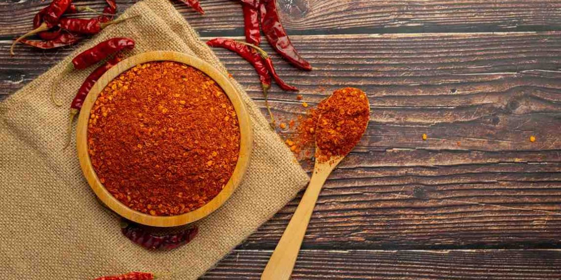 Bowl of chili powder with dried peppers on wooden table.