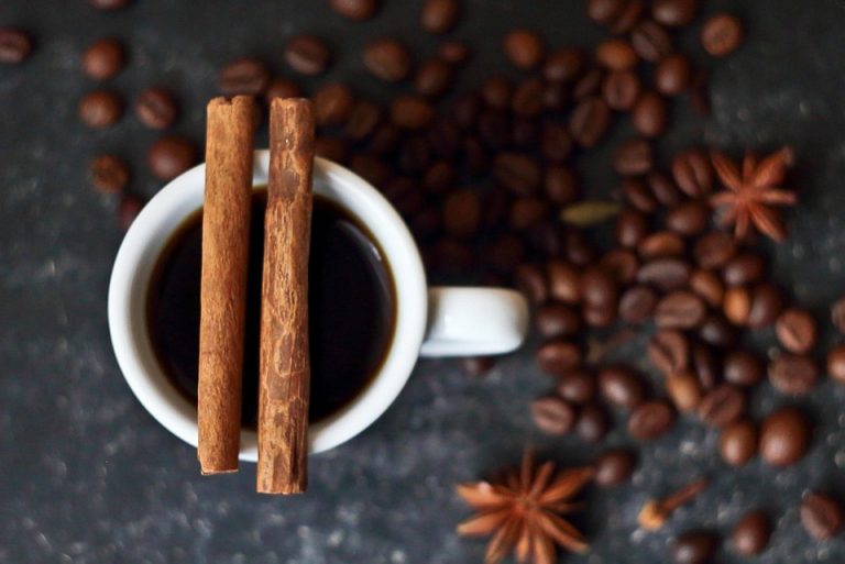 Cup of coffee with cinnamon sticks surrounded by coffee beans.