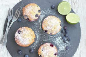 Blueberry muffins with powdered sugar on slate, limes and blueberries on side.