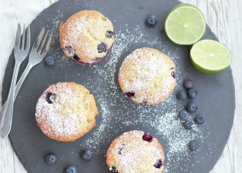 Blueberry muffins with powdered sugar on slate, limes and blueberries on side.