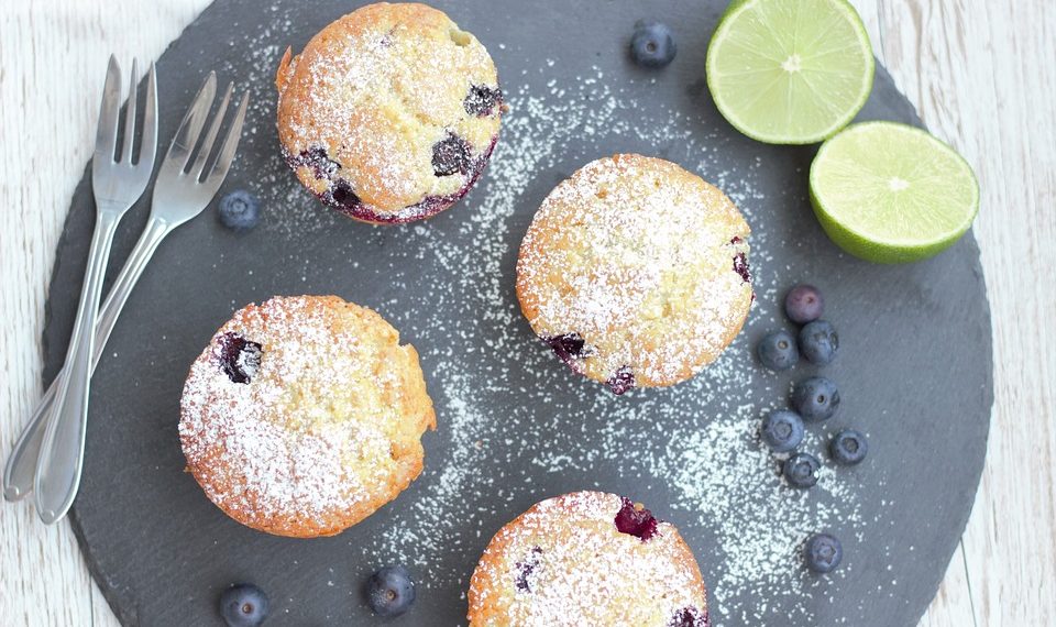 Blueberry muffins with powdered sugar on slate, limes and blueberries on side.