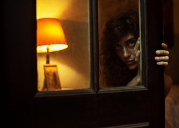 Woman peering through dimly lit doorway with lamp in background.