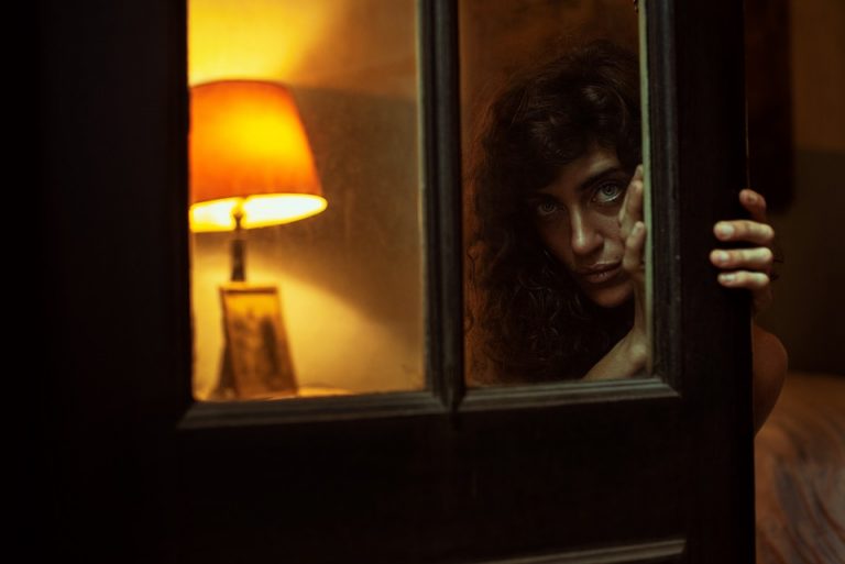 Woman peering through dimly lit doorway with lamp in background.