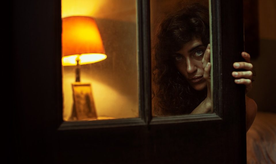 Woman peering through dimly lit doorway with lamp in background.