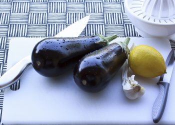 Eggplants, lemon, and garlic on a cutting board with knives.