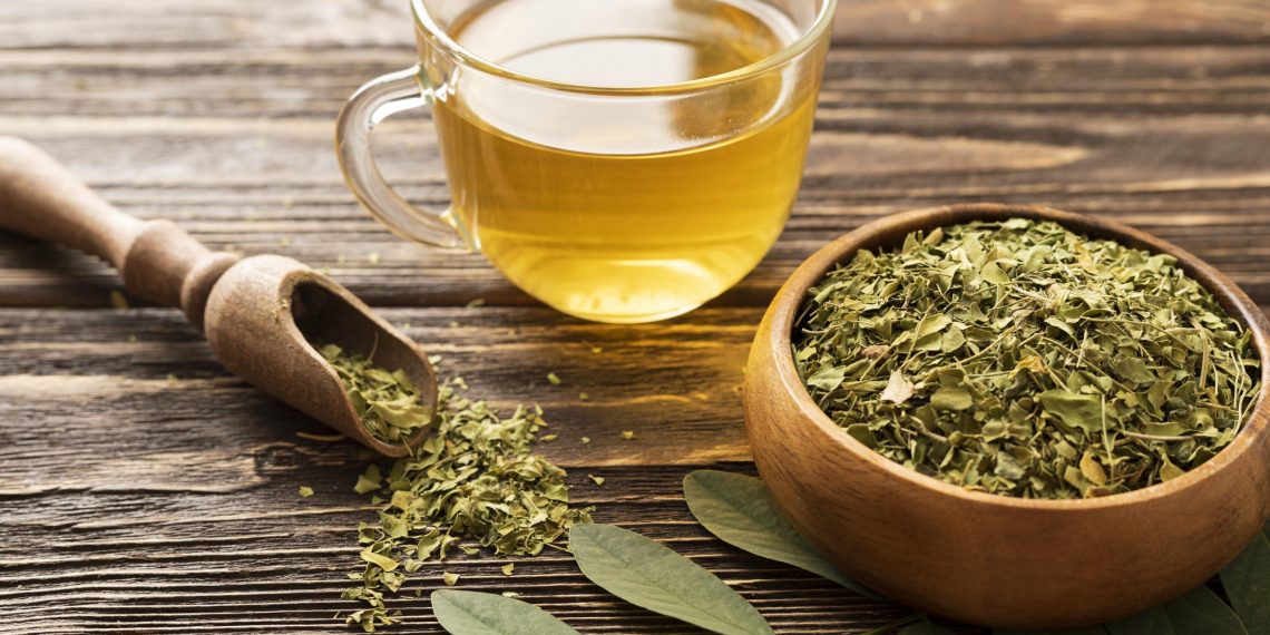 Cup of moringa tea next to dried moringa leaves in a wooden bowl.