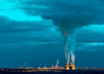 Power plant emitting smoke under cloudy sky with blue hues.