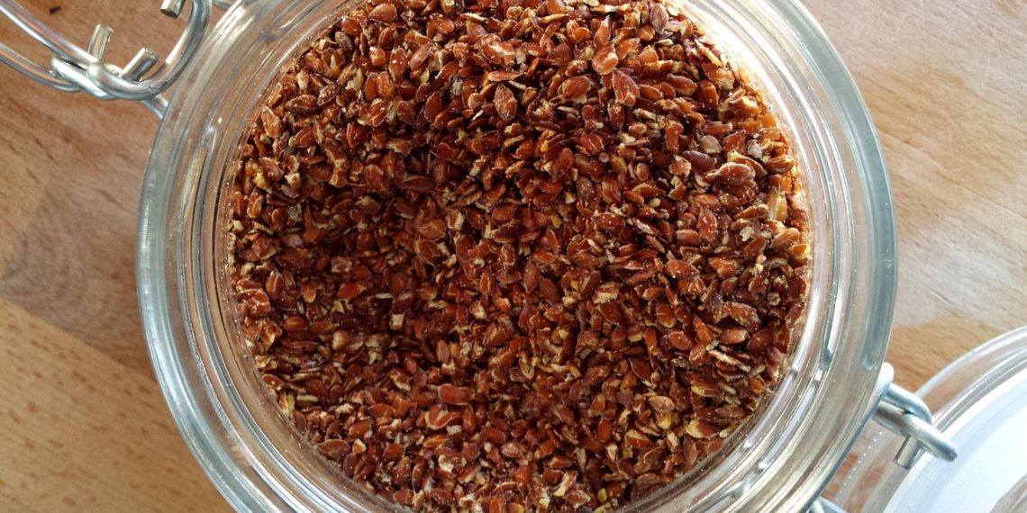 Jar filled with brown flaxseeds on wooden table top.