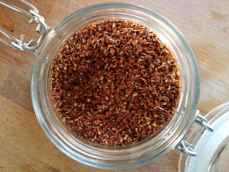 Jar filled with brown flaxseeds on wooden table top.