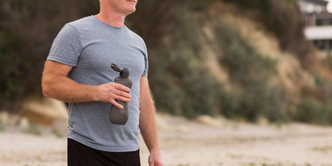 Man holding water bottle on the beach in athletic attire.
