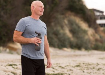 Man holding water bottle on the beach in athletic attire.
