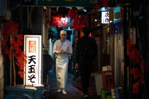 Chef walks through narrow Tokyo alleyway illuminated by lanterns.