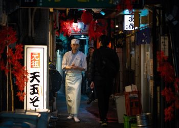 Chef walks through narrow Tokyo alleyway illuminated by lanterns.