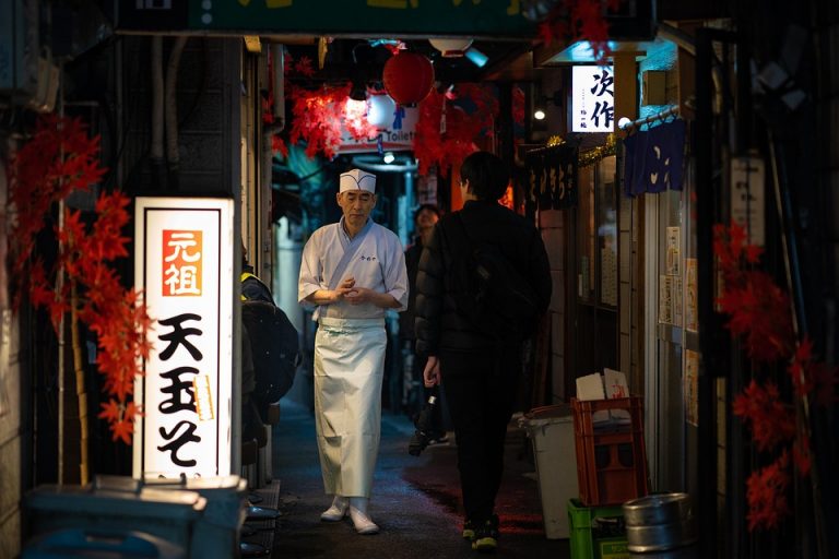 Chef walks through narrow Tokyo alleyway illuminated by lanterns.