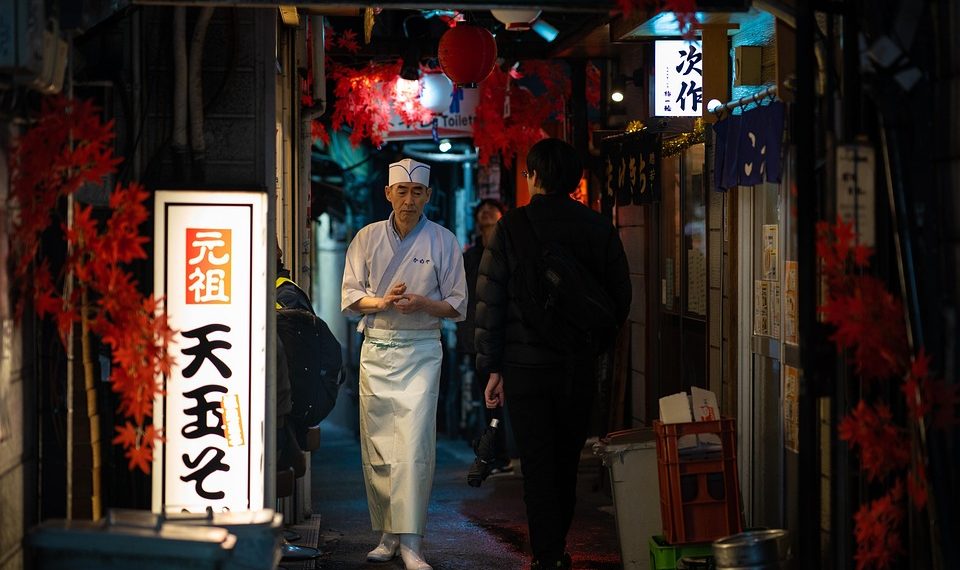 Chef walks through narrow Tokyo alleyway illuminated by lanterns.