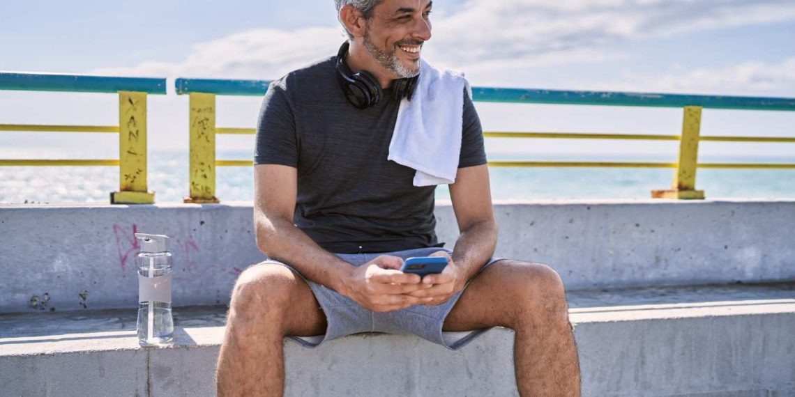 Man relaxing on bench by the beach, smiling while using smartphone with headphones and water bottle nearby.