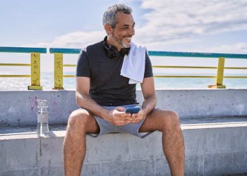 Man relaxing on bench by the beach, smiling while using smartphone with headphones and water bottle nearby.