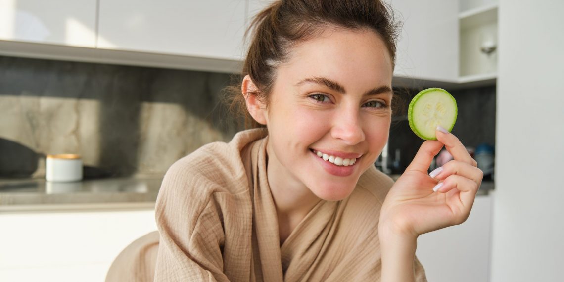 Woman smiling with cucumber slice in kitchen.