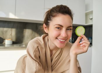 Woman smiling with cucumber slice in kitchen.