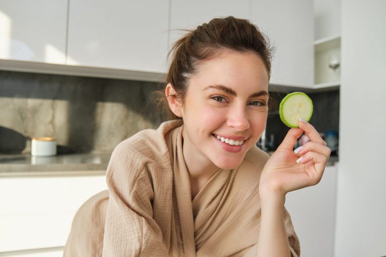 Woman smiling with cucumber slice in kitchen.