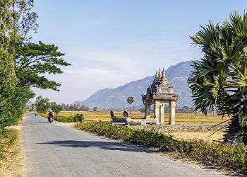 Rural road leading to traditional Cambodian gate, with distant mountains.