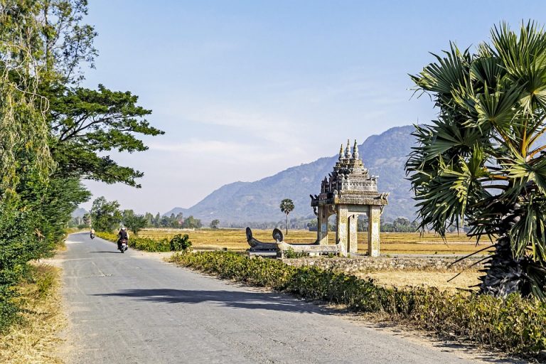 Rural road leading to traditional Cambodian gate, with distant mountains.