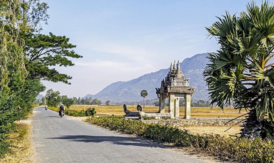 Rural road leading to traditional Cambodian gate, with distant mountains.