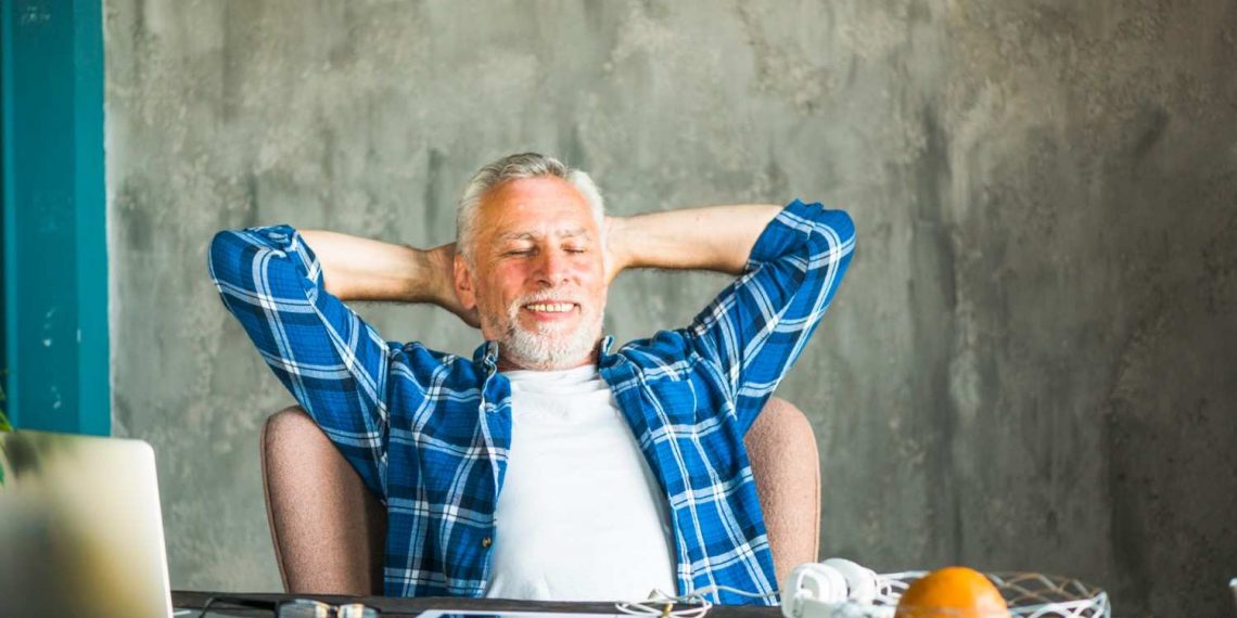Man relaxing at desk with laptop, wearing blue plaid shirt.