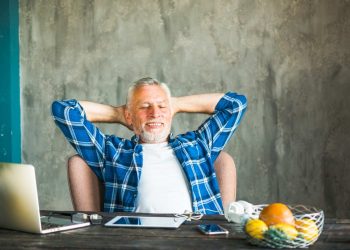 Man relaxing at desk with laptop, wearing blue plaid shirt.