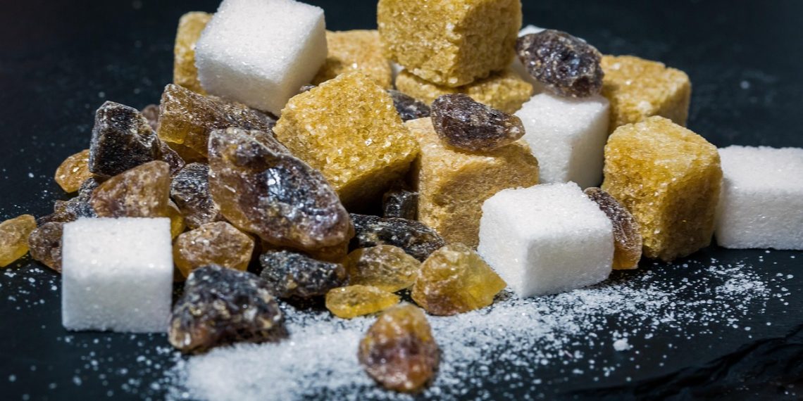 A mix of brown and white sugar cubes alongside raw amber and dark sugar crystals on a dark surface.