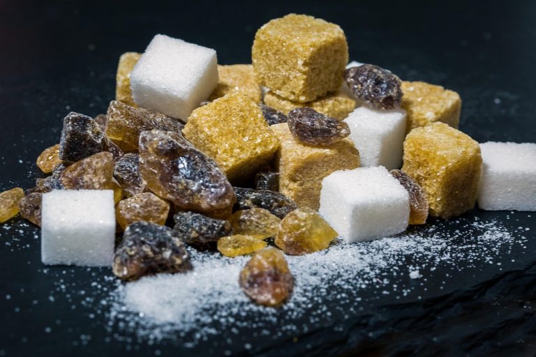 A mix of brown and white sugar cubes alongside raw amber and dark sugar crystals on a dark surface.