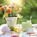 Flowers in a pot with tea, cupcake, and lemons on a floral tablecloth in sunlight.