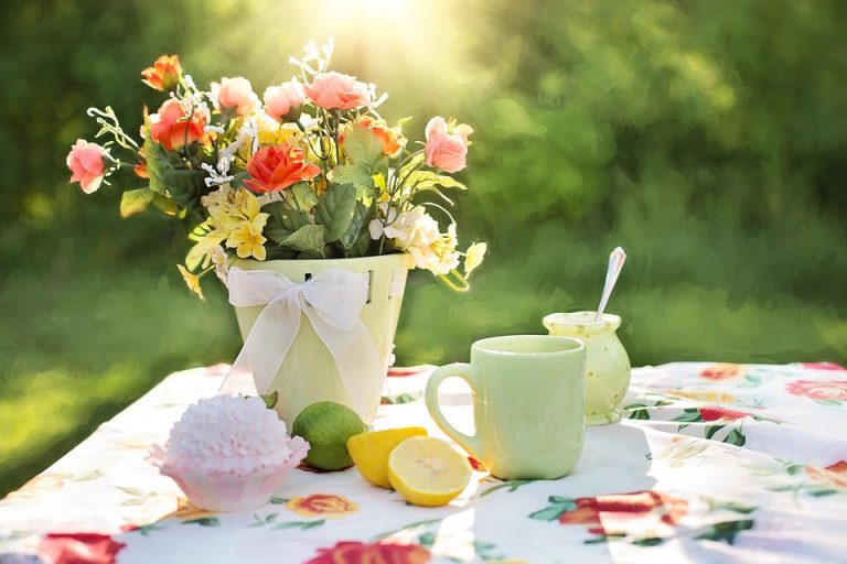 Flowers in a pot with tea, cupcake, and lemons on a floral tablecloth in sunlight.
