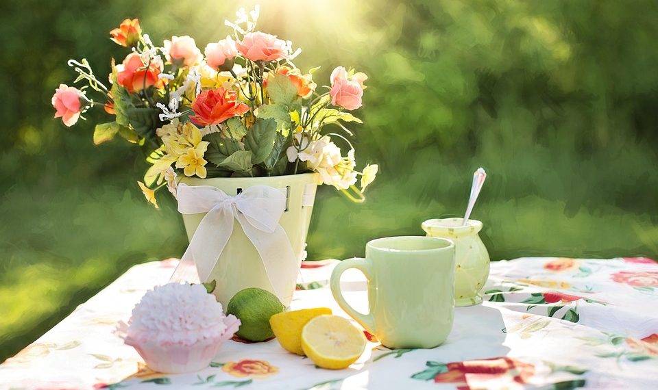 Flowers in a pot with tea, cupcake, and lemons on a floral tablecloth in sunlight.