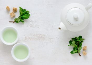 White teapot and cups with mint leaves and sugar cubes on a table.
