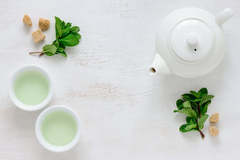 White teapot and cups with mint leaves and sugar cubes on a table.
